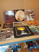 Photo of 9 vinyl records arranged on a table with visible album covers showing portraits, abstract art, and bright colors.