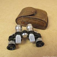 Photo of the pair of black and silver Baker Terra opera binoculars alongside the brown leather case.