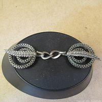 Top view of the vintage silver tone cape hook on a black display, showing both circular filigree discs connected by a curved link and two textured leaf-like pins passing through each disc.