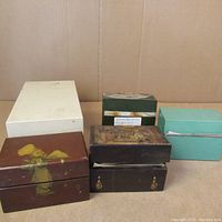 Five vintage recipe boxes of varying materials and styles arranged against a plain backdrop. Boxes include wood with painted art, metal with label, and colored cardboard types.
