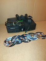 Original Xbox console with attached power cable, two wired controllers placed on top and six discs arranged in front on a cardboard background.