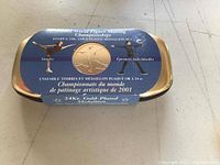 Front view of unopened rectangular metal presentation case with label showing gold-plated medallion and event details.