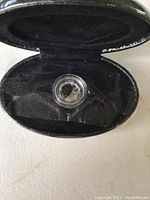 Black oval hinged case, interior view showing quarter coin inside protective clear capsule, resting on black velvet lining.