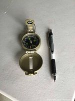 Top-down photo of open brass-colored vintage compass next to a pen for size reference, showing the compass dial with directional markings and sighting window in the lid.