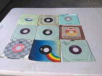 Nine 45 RPM vinyl records laid out in three rows of three on a table, showing the front covers with various colors and label designs.