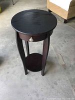 Full view of the dark wood round plant stand showing the top, legs, small drawer with knob, and lower shelf.