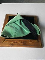 Green Robin Hood hat with black feather shown from left side, placed on black velvet surface with wooden frame around.