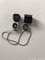 Two vintage items: a Soligor Auto Light Meter with black leather carrying case and string strap, and a camera lens inside a black and red stitched protective case, both made in Japan.