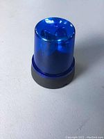 Close-up photo of blue flashing emergency rotating light from an angle showing its blue plastic cover and black base on a plain surface.