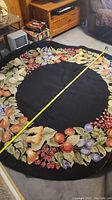 Photo showing the full circular rug with a tape measure across its diameter, featuring black center and multi-colored fruit and leaf border.