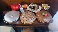 Full view of all items on wooden table including metal and ceramic pie plates, decorative plates, ceramic cat plates, metal apple container, character mug, and cake slice server.