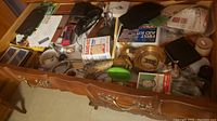 Top-down view of an open wooden drawer filled with assorted small items including wallets, watches, and household goods.