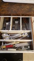 Drawer with assorted metal forks, spoons, and knives, plus several kitchen hand tools such as can opener and peeler.