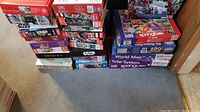 Photo showing a large stacked assortment of jigsaw puzzle boxes of various themes and piece counts on carpeted floor.