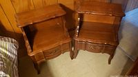 Two matching wooden bedside tables with open shelves, drawers, and scalloped edges.