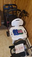 Wide shot showing white plastic shower chair with backrest and six dumbbells on the seat, two walkers in background, and part of elliptical machine below chair