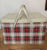 Front view of large vintage metal picnic basket showing plaid exterior and white textured lid with dual handles folded up.