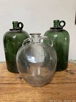 Three large glass bottles: two green bottles with loop handles near the neck and one clear glass bottle with dual handles and textured decorative details.