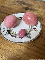 Three pink marble eggs arranged on the decorative saucer showing the different sizes and glossy finish of the eggs and details of the saucer's illustrations.