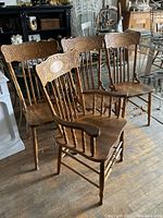 Four solid wood pressback chairs grouped together showing carved decorative top rails, spindled backs, and contoured wooden seats. Two chairs have arms, two are armless. Good used condition.
