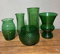 Five vintage green glass vases with various shapes and textures arranged side by side on a wooden surface.