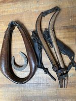 Photo showing two antique horse collars, a metal horseshoe, and black leather harness straps with metal rings and buckles, all laid out on a wooden surface.
