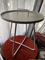Full view of the painted white metal side table showing round top with handle and triangular tubular legs.