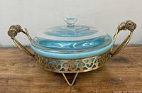 Side view of turquoise glass bowl with clear lid sitting in gold-toned metal stand with decorative cut-out pattern and wire wrapped handles.