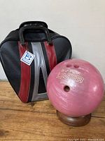 Photo of vintage pink Brunswick Laser bowling ball next to black, red and silver striped zippered carrying bag with handle and branded tag.