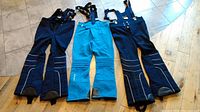 Three pairs of men's ski pants on floor - two navy blue and one turquoise, with suspenders attached