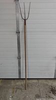 Full view of the antique hay fork standing upright against a garage door, showing the entire length and metal fork top.