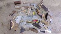 Overview of various masonry tools spread out on a concrete floor including trowels, scrapers, saws, and brushes.