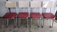Front view of four matching mid-1900s chairs with red vinyl seats and beige backrests showing wear.
