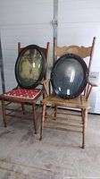 Wide view showing two vintage wooden pressback chairs. One with red patterned cushion and the other with carved seat, each holding an oval photo frame with curved glass.