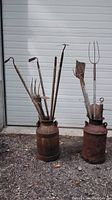 Two rusty metal milk cans filled with various long-handled rusty farm tools against a white garage door.
