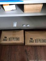 Four cardboard boxes labeled 'Jiale Premium Quality Bed Pillows' stacked under a shelf on a wooden floor