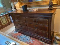 Full view of wooden dresser showing six drawers and three smaller drawers on top, placed against a wall on a patterned rug. Two lamps and other household items on top of dresser.