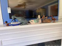 Wide photo showing all six duck figurines of varying materials and the model ship arranged on a white mantle with a TV behind them.