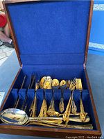 Full view of gold plated flatware and serving utensils arranged in blue velvet-lined wooden presentation box showing spoons, forks, and serving pieces.