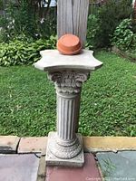 Photo of concrete statue pedestal column with ornate carved top and base, holding one upside-down plastic flower pot, set on a flagstone patio with grass in background.