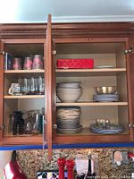Kitchen cabinet with glassware, cups, and plates. Top left shows several clear glasses and colored cups. Bottom left has clear glasses and some dark-colored tall glasses. Center and right side include white bowls and plates, and blue decorative plates with floral patterns.
