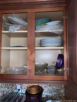 Kitchen cabinet with ceramic plates, bowls wrapped in bubble wrap on top shelves, clear glass bowls, plastic containers with lids, and metal trays on bottom shelf.