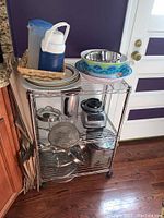 Wide view of rolling metal shelf loaded with kitchenware including blender, bowls, trays, jugs, plates, and containers.