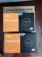 Three boxed items arranged on floor: one box labeled Crystal Bedside Lamp and two boxes of Twinkle Star Curtain String Lights 300 LED.