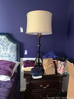 Full view of the tall table lamp with beige fabric shade next to a bed and folded clothes around the base.