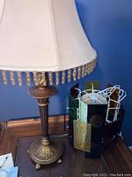 Full view of antique-style bronze table lamp with cream fabric shade with beaded fringe on wooden floor against blue wall.