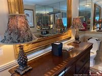 Pair of pineapple table lamps placed on a wooden dresser with gold-framed mirrors in the background.