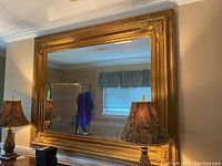 Full view of the large ornate gold-framed mirror hanging on the wall with lamps reflected in the glass, showing size and placement.