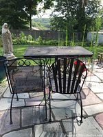 Photo of black metal mesh patio table and two of the four matching chairs with different backrest designs outdoors on stone patio surface surrounded by garden and trees.