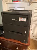 Photo showing Brother HL8180DW black laser printer with dual lower paper trays on wooden surface.